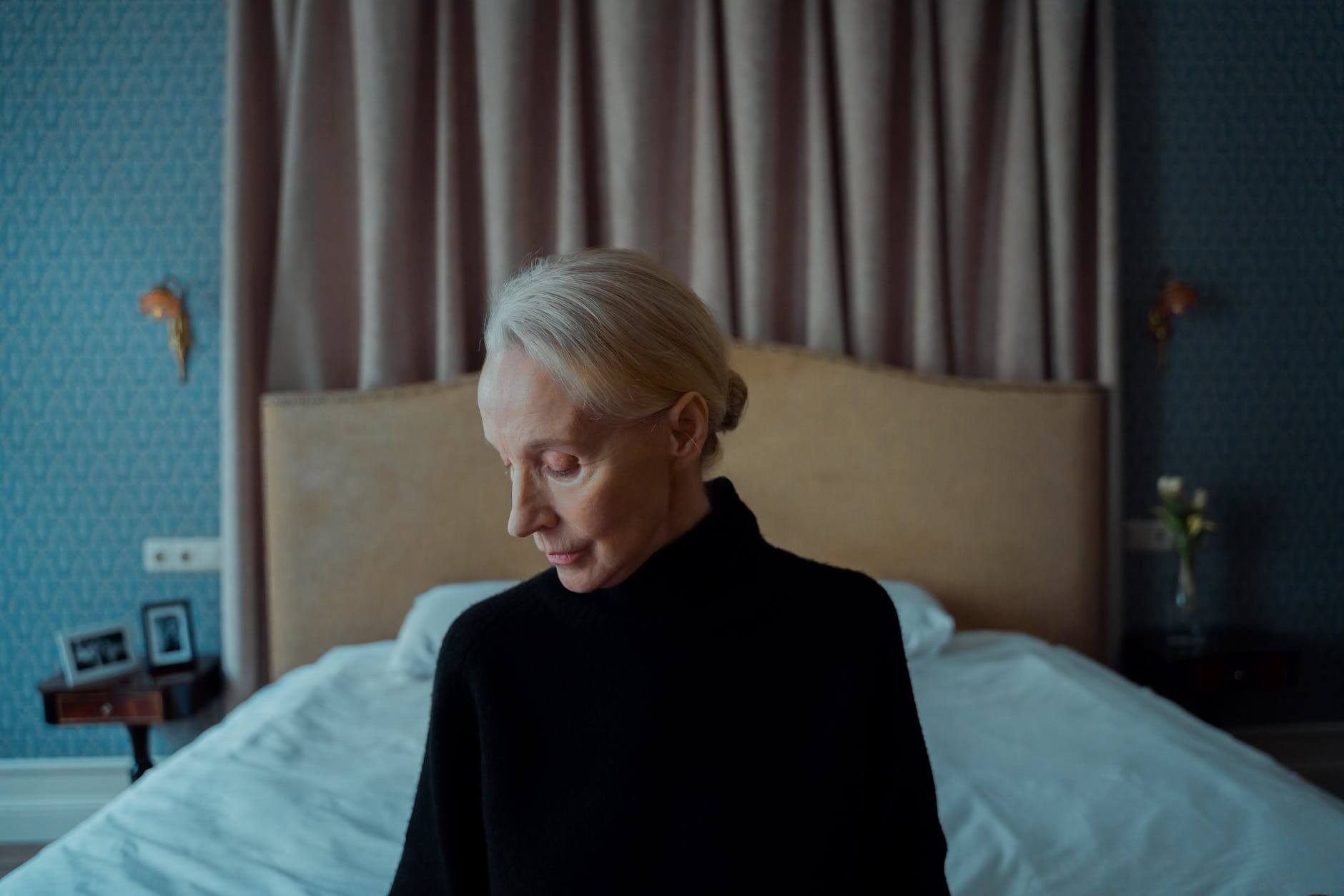 elderly woman in black dress sitting on foot of the bed feeling sad